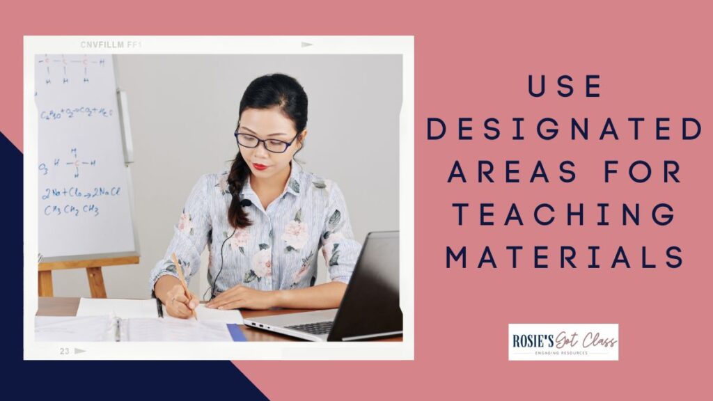 Teacher at her desk with an open laptop and writing on papers with an easel with writing on it in the background that says use designated areas for teaching materials